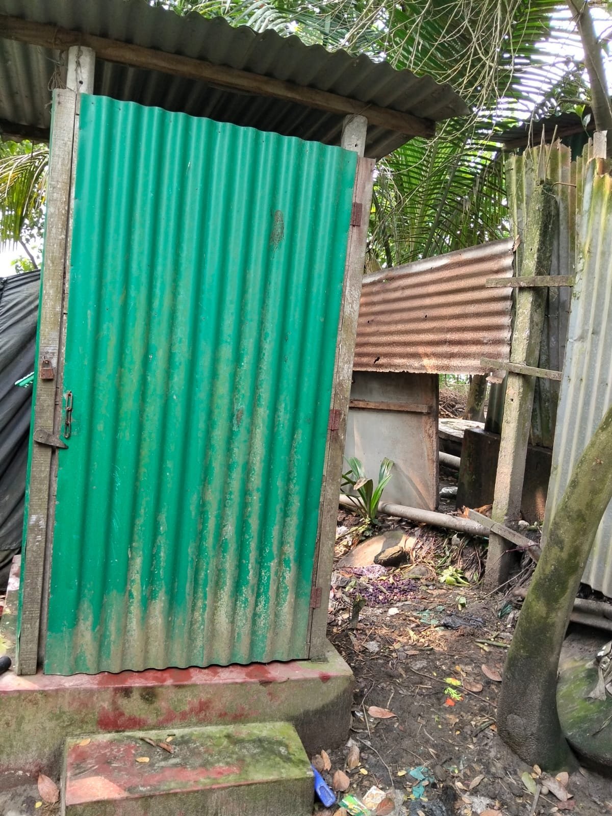Green tin latrine with a chain latch on the door, a rusted tin fence behind.